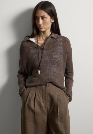 Junge Frau mit langem dunklem Haar trägt einen lockeren braunen Strickpullover, passende hochtaillierte Faltenhose und eine schwarze Halskette mit Anhänger, Hände in den Taschen.