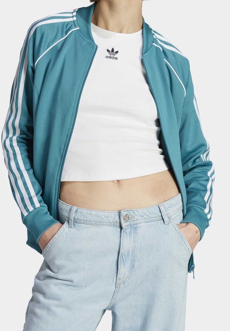 Tyrkysová sportovní bunda s bílými pruhy podél rukávů, v kombinaci s bílým vroubkovaným crop topem a světle modrými džíny s vysokým pasem.