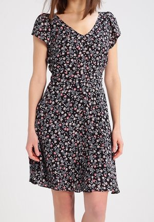 Femme portant une robe noire à manches courtes avec de petits motifs floraux roses et blancs et un décolleté en V, debout devant un fond uni.