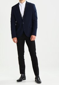 Marinblå blazer med spårad slag, enkel knapp och två framfickor, i kombination med en vit skjorta och svarta slim fit-byxor.