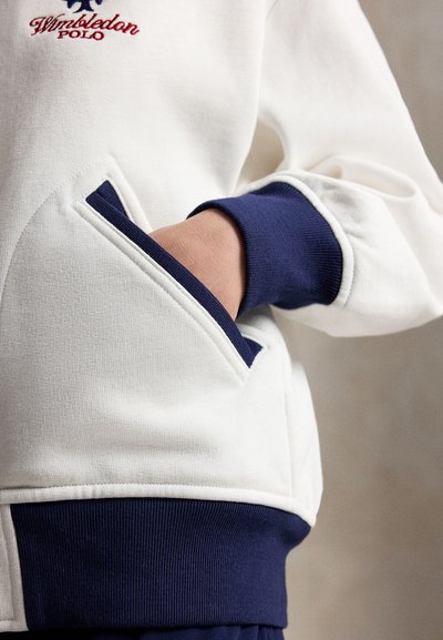 Hand wearing white Wimbledon Polo sweatshirt with blue ribbed cuff and pocket trim, hand inserted in side pocket.