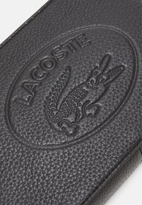 Portefeuille en cuir noir texturé, mettant en avant un logo crocodile Lacoste en relief et un détail de marquage circulaire. Fini sans couture et sans quincaillerie visible.