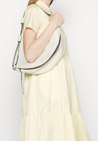Femme en robe jaune clair tenant un grand sac à bandoulière en cuir blanc en forme de croissant avec une fermeture éclair noire.