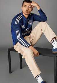 In een lange blauwe trui met witte strepen, beige broek met drukknopen en blauwe sneakers. Model zit op een zwarte bank.