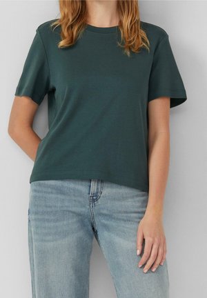 Kvinde med lys hud og rødt hår iført en mørkegrøn kortærmet t-shirt og lyseblå jeans, stående foran en ensfarvet lys baggrund.