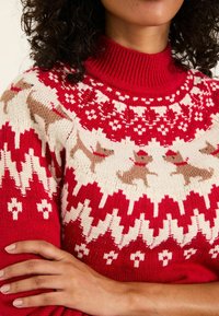 Frau in einem roten und cremefarbenen Strickpullover mit Rentieren und geometrischen Mustern, Arme verschränkt, lockiges, dunkles Haar teilweise sichtbar.