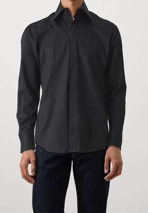 Man draagt een zwarte blouse met lange mouwen en verborgen knoopsluiting en donkerblauwe spijkerbroek, staande voor een effen achtergrond.