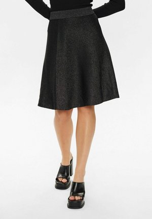 Jupe évasée noire avec un éclat métallique. Texture lisse, large ceinture, longueur genou, associée à des chaussures à plateforme noires.