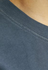 Tums zilā kokvilnas T-krekls tuvu, ar ribotu apaļu kakla izgriezumu un smalku tekstūru, izceļot vīles gar malu.