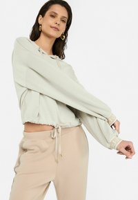 Sudadera cortada de color beige claro con un dobladillo ajustable, mangas largas y acentos de logo en los puños, combinada con joggers beige. Textura suave.