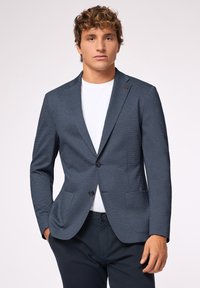 Granatowa marynarka z teksturowanej tkaniny, z zapięciem na jeden guzik i dwoma przednimi kieszeniami. Nosi się ją na białej koszulce z czerwonymi akcentami.