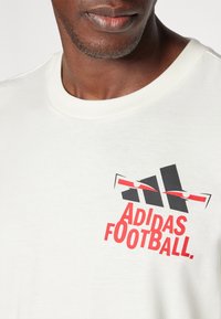 Hvid bomulds t-shirt med sort og rød "ADIDAS FOOTBALL" grafiktryk. Korte ærmer og klassisk rund halsudskæring.