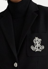 Černý blazer s jemnou texturou, vybavený bílým vyšitým logem na levé kapse a stříbrným detailem knoflíku na přední straně.