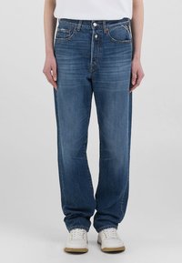 Jeans in denim blu con una vestibilità rilassata, dotati di cinque tasche, chiusura con bottone e cuciture in contrasto. Indossati con sneaker bianche.