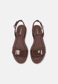 Brune flate sandaler med bred rem, metallisk detalj, justerbar ankelstropp og glatt teksturert såle, med logo på sålen.