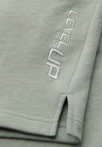 Tissu vert clair avec "LEVEL UP" et "OMEGA" brodés en blanc près d'une petite fente latérale sur l'ourlet.