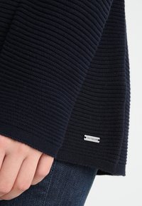 Granatowy sweter wykonany z prążkowanego dzianiny, z fakturowanym wzorem i metką z napisem "TOM TAILOR" na dolnym brzegu. Palce chwytają brzeg.