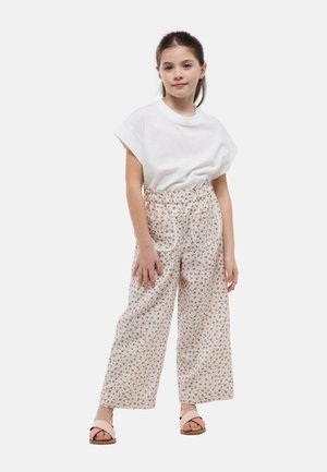 Une fille tient debout portant une chemise blanche à manches courtes, un pantalon large taille haute à motifs floraux, et des sandales beiges à brides croisées sur un fond blanc uni.