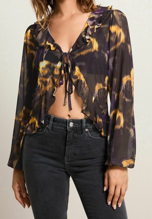 Kvinde iført gennemsigtig langærmet blomstret bluse med flæsekanter bundet foran, kombineret med sorte højttaljede jeans.