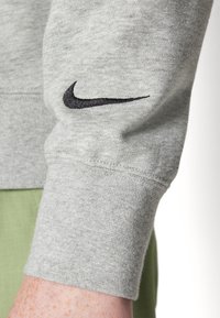 Grå sweatshirt med en svart Nike-logotyp på ärmslutet. Tyget verkar mjukt med en slät textur, och ärmen har en ribbad avslutning.