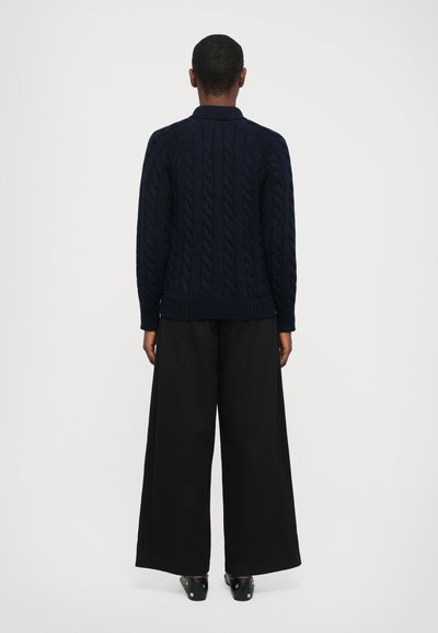 Pull en maille torsadée bleu marine avec un col et un ourlet côtelés, associé à un pantalon noir à jambes larges. La tenue présente un design texturé en tricot.