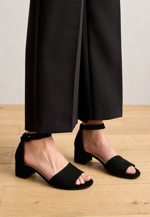 Zwarte suede open hakken met een blokhak en enkelbandje. Het ontwerp heeft een zachte textuur en een minimalistische stijl.
