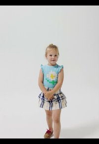 Jeune fille souriante, portant un haut sans manches bleu avec un motif de fleur, une jupe à carreaux et des chaussures marron avec des lacets rouges.