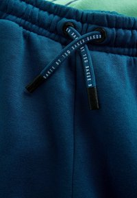 Niebieskie dresy mają elastyczny pas z czarnymi akcentami. Materiał jest miękki, o gładkiej teksturze i jednolitym kolorze. Widoczna metka marki.