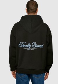 Svart oversized hettegenser med snøring i hetten, med lys blå grafisk tekst på baksiden som sier "Eternally Blessed."