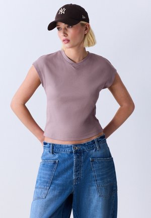 Kobieta w bezrękawniku w kolorze mauve, niebieskich szerokich dżinsach z dużymi przednimi kieszeniami i czarnej czapce z daszkiem, patrząca na bok.