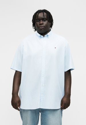 Άνδρας με σκουρόχρωμο δέρμα και dreadlocks, ντυμένος με ανοιχτό μπλε πουκάμισο με κοντά μανίκια και μπλε τζιν, στέκεται μπροστά σε απλό φόντο.