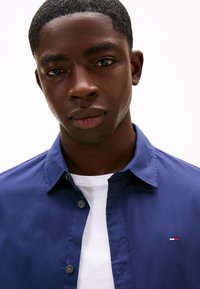 Camisa de botão azul-marinho com um logótipo subtil no peito esquerdo, usada sobre uma t-shirt branca. Textura suave, design clássico de colarinho.