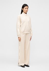 Jakna z zadrgo v kremni barvi s stranskim logotipom, kombinirana z ujemajočimi se širokimi hlačami. Gladek tekstura, minimalističen dizajn in sproščen kroj.