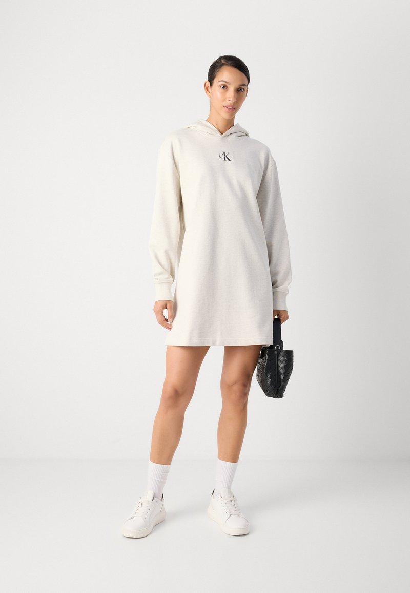 Hooded jurk van lichtbeige stof, lange mouwen, met een centraal logo, gecombineerd met witte sneakers en een kleine zwarte gevlochten handtas.