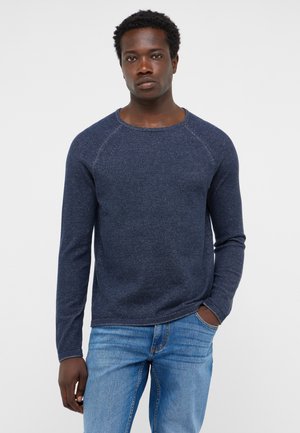 Mann mit kurzen lockigen Haaren trägt einen dunkelblauen Langarmpullover und hellblaue Jeans, steht mit einer Hand in der Tasche vor weißem Hintergrund.