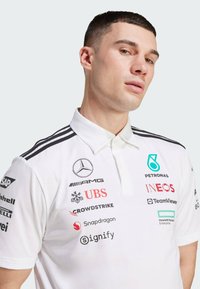 White polo shirt with a collared design, featuring multiple sponsor logos in various colors and black stripes on the shoulders.