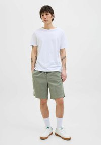 Camiseta blanca de algodón combinada con pantalones cortos verde oliva. El modelo lleva zapatillas blancas con suelas de goma y calcetines blancos. Diseño liso y casual.