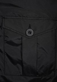 Černá látka s hladkým povrchem má výraznou náprsní kapsu se zapínáním na patentku a vyztuženým švem.