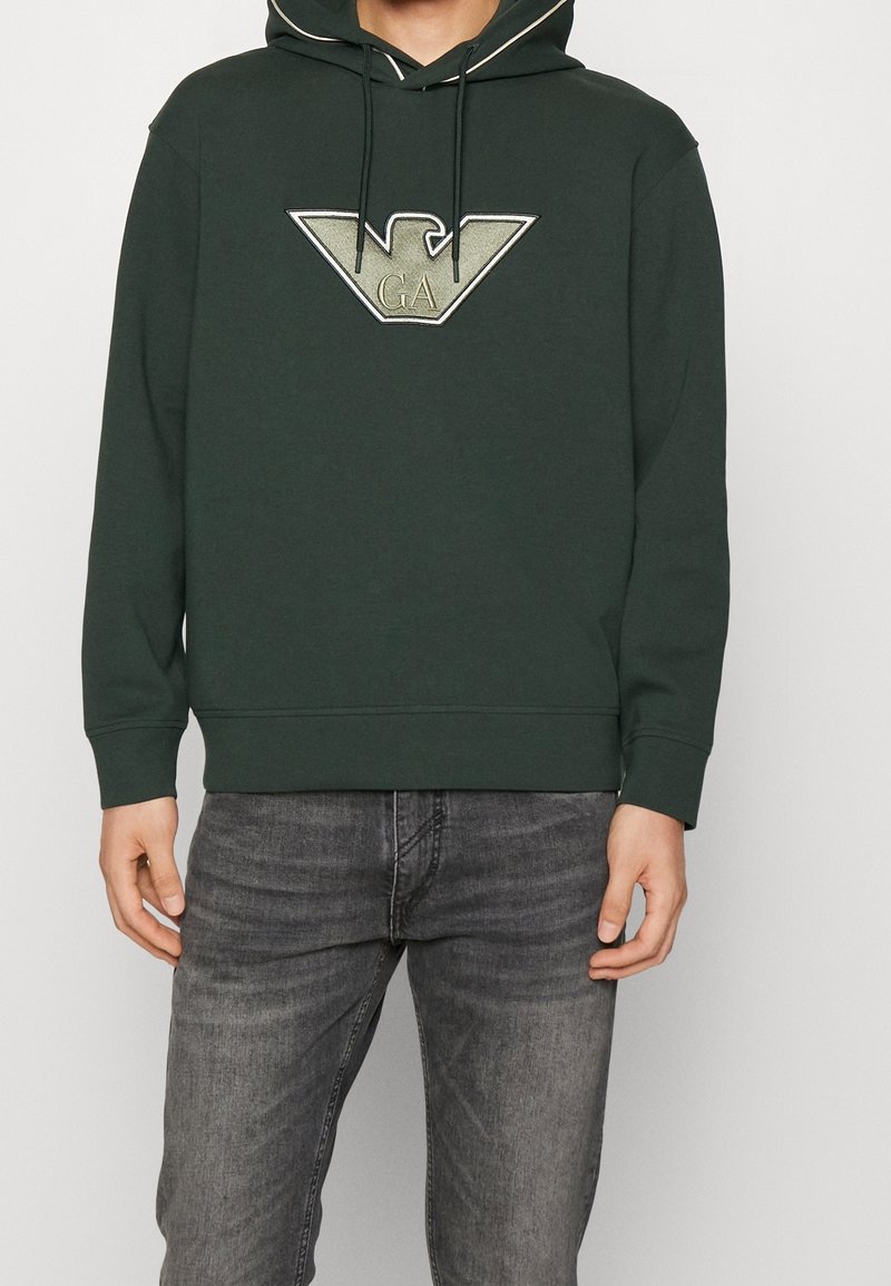 Mörkgrön hoodie med en känguruficka, skosnören i huvan och en stor metallisk logotyp på bröstet. Mjuk textur och ribbade ärmslut.