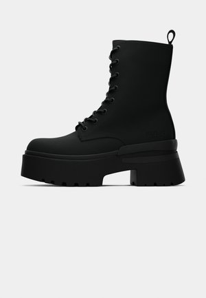 Bota de combate preta com atacadores, sola plataforma grossa, salto robusto e laço na parte de trás, apresentada de lado sobre um fundo branco.