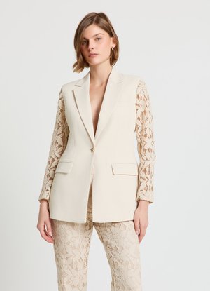Blazer beige con maniche in pizzo, caratterizzato da una profonda scollatura a V, vestibilità strutturata e due tasche frontali. Abbinato a pantaloni in pizzo tono su tono.