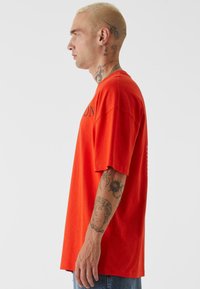 Rød oversized bomullst-skjorte med kortermer, med svart tekst på forsiden og baksiden. Synlige tatoveringer på modellens arm.