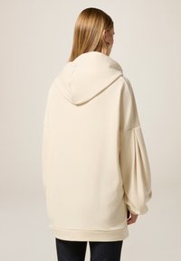 Hoodie oversize crème avec une grande capuche, des épaules tombantes et des manches bouffantes. L'ourlet contrastant complète le design décontracté.