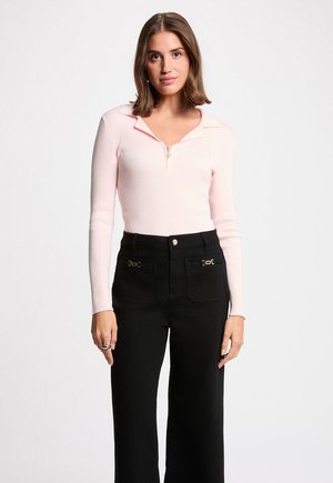 Vrouw met halflang bruin haar, gekleed in een lichtroze top met lange mouwen en een rits aan de voorkant, en zwarte hoge taillebroek met zakdetails.