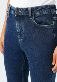 Tmavě modré džíny z denimu s hladkou texturou, s knoflíkovým zapínáním, klasickým pětikapsovým designem a kontrastním prošíváním.