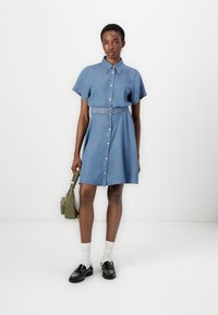 Denim shirt dress with short sleeves, button front, and belted waist. Light blue color, knee-length, worn with black loafers and a green bag.