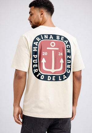 Man wearing beige t-shirt with large anchor graphic and text "Marina Beach Puerto de la Cruz 2024" on the back.