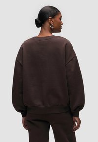 Felpa oversized marrone con collo a giro, spalle scese e polsini a costine. Tessuto morbido e vestibilità rilassata, abbinata a pantaloni coordinati.