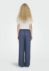 Pantalon en lin bleu à jambes larges avec une taille élastique, associé à un t-shirt blanc uni, vu de dos. Cheveux longs, texturés et bouclés visibles.