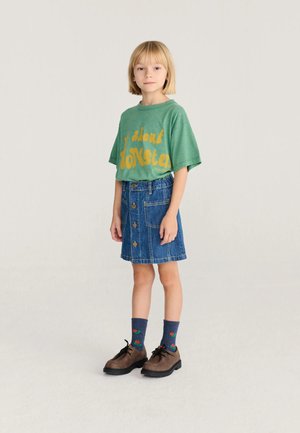 Enfant aux cheveux blonds courts portant un t-shirt vert avec un texte jaune, une jupe en jean avec des boutons, des chaussettes à motifs et des chaussures marron, debout sur un fond uni.